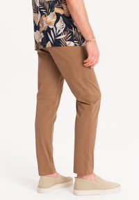 Pantaloni chinos beige con una vestibilità slim e una texture liscia, abbinati a scarpe slip-on beige. La camicia presenta un motivo floreale scuro.