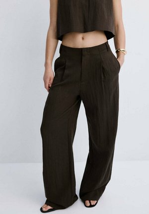 Woman wearing loose-fitting dark brown high-waist trousers, cropped sleeveless top, sandals, and gold bracelet with hands in pockets.