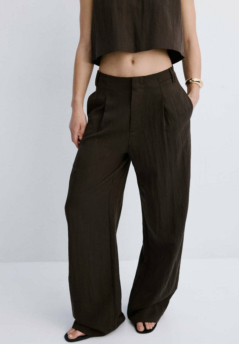 Femme portant un pantalon ample taille haute marron foncé, un top court sans manches, des sandales et un bracelet en or, les mains dans les poches.