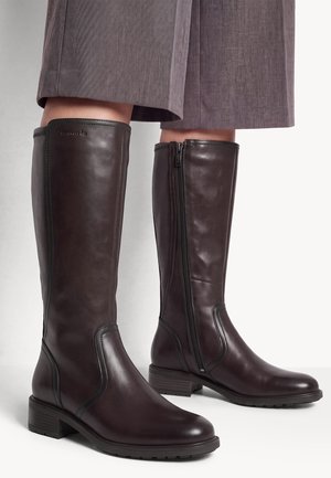 Bottes montantes en cuir marron avec fermeture éclair latérale portées avec un pantalon gris court, montrant une finition lisse et des talons bas empilés.