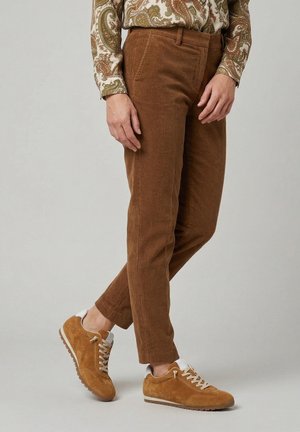 Osoba ubrana w brązowe spodnie sztruksowe, koszulę z długim rękawem w wzory oraz jasnobrązowe zamszowe trampki z białymi sznurówkami i podeszwami.