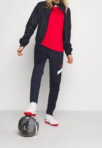 Nike träningsoutfit bestående av en röd tröja och svart jacka, tillsammans med marinblå byxor. Vita och röda fotbollsskor, svart fotboll på marken.