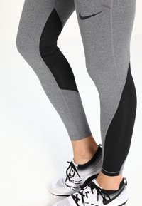 Leggings de sport gris avec des accents de panneaux noirs, présentant une coupe ajustée et une texture lisse. Associés à des chaussures de sport noir et blanc.