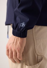 Navy blue jacket made of lightweight fabric, featuring elastic cuffs and a subtle embroidered logo on the sleeve. Close-up of a hand.