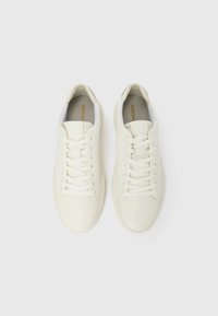 Paire de baskets basses blanches avec lacets, surface texturée et nom de la marque visible sur la semelle intérieure, présentées sur un fond clair uni.