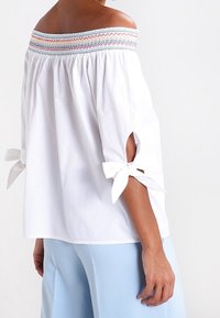 Donna che indossa una blusa bianca off-shoulder con scollatura smock multicolore e maniche con fiocco bianco, abbinata a pantaloni azzurri.