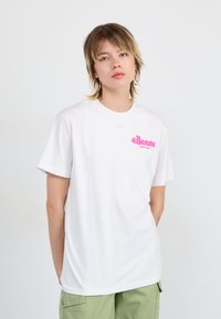 T-shirt en coton blanc avec un logo ellesse rose sur la poitrine. Manches courtes avec un col rond, texture lisse. Porté avec un short cargo vert.