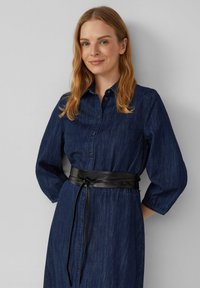 Vrouw met schouderlang blond haar, gekleed in een donkerblauwe denim knoopjurk met driekwart mouwen en een zwarte leren tailleband.