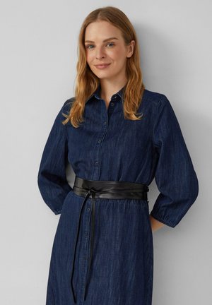 Frau mit schulterlangem blonden Haar, die ein dunkelblaues Jeanshemdkleid mit 3/4-Ärmeln und einem schwarzen Ledergürtel trägt.