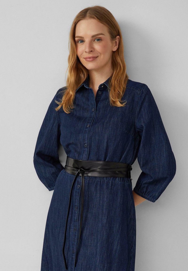 Vrouw met schouderlang blond haar, gekleed in een donkerblauwe denim knoopjurk met driekwart mouwen en een zwarte leren tailleband.