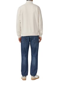 Helle beiger Sweatshirt mit hohem Kragen, kombiniert mit locker sitzenden dunkelblauen Jeans und weißen Sneakers mit beigen Akzenten.