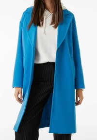 Cappotto blu, oversize, con rever a punta, chiusura con un solo bottone e due tasche laterali. Indossato sopra un hoodie bianco e pantaloni a righe.
