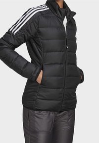 Černá péřovka s vertikálním švem, vysokým límcem, zipovým zapínáním a třemi bílými pruhy na ramenou. Texturovaný, vycpaný materiál.