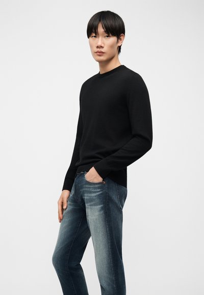 Jeune homme debout, une main dans la poche, portant une chemise noire à manches longues et un jean bleu délavé, sur un fond blanc uni.