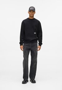 Must mustaga ümarate kaelustega ning väikese logoga, koos tumedate hallide teksadega. Mudel kannab halli mütsi ja musti jalatseid.