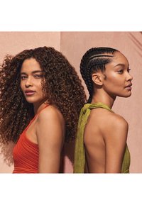 Se presentan dos mujeres, una con una blusa texturizada de color óxido y la otra con un top halter verde liso, frente a una pared rosa.