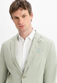 Helle grüne Leinenjacke mit einem Knopf, Reverskragen und einer Brusttasche, in der ein blauer, gestreifter Taschentuch steckt, getragen über einem weißen Hemd.