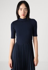 Jūras kara flotes ribotas kleitas ar augstu kakla izgriezumu un īsām piedurknēm, ar plisētu svārku un gludu tekstūru. Minimalistisks dizains ar plānām detaļām.