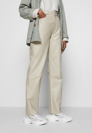 Persona vistiendo pantalones beige de pierna ancha, zapatillas blancas y grises, una camisa a rayas y un blazer verde a cuadros con una mano en el bolsillo.