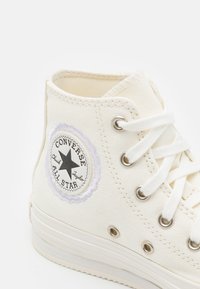 Zapatilla alta de lona blanca con ojales plateados, cordones blancos y un parche circular en blanco y negro con un estrella.