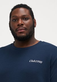 Marineblaues Sweatshirt mit Rundhalsausschnitt. Verfügt über weißen, auf der linken Brust gestickten Text "Club1990". Der Stoff erscheint weich und glatt.