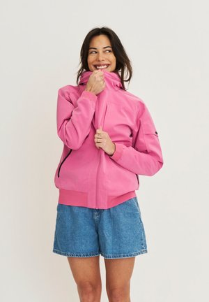 Femme souriante en train de fermer une veste rose vif, portant un short en jean bleu, debout devant un fond clair uni.
