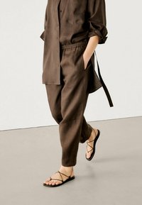 Set van een bruine linnen shirt en broek met een relaxte pasvorm, knoopsluiting en opgerolde mouwen, gecombineerd met zwarte sandalen met bandjes.