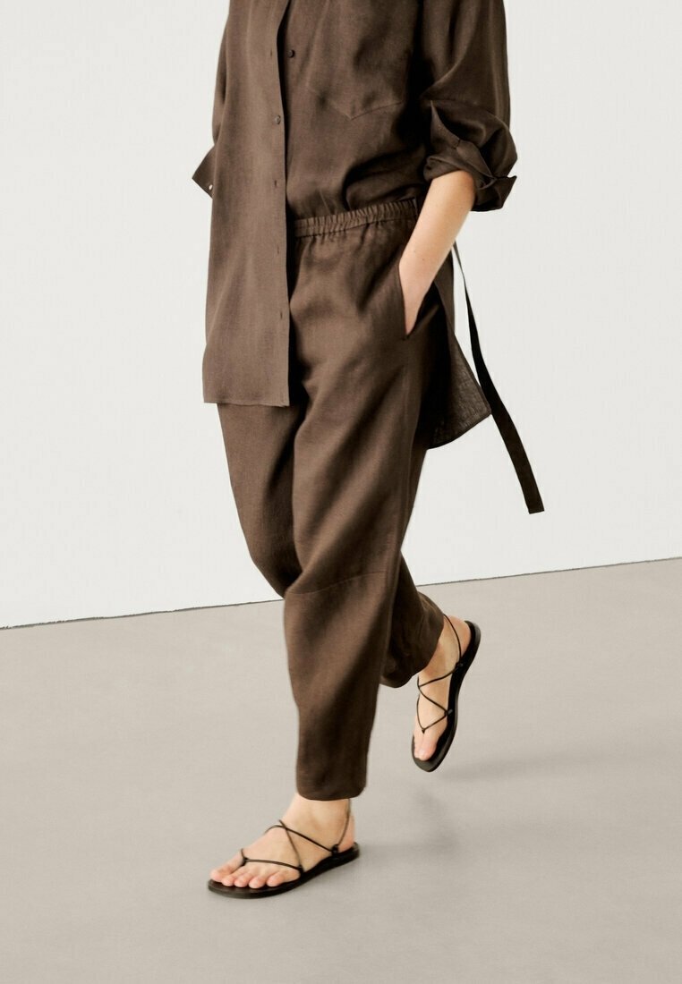 Set van een bruine linnen shirt en broek met een relaxte pasvorm, knoopsluiting en opgerolde mouwen, gecombineerd met zwarte sandalen met bandjes.