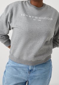 Harmaa huppari, jossa on pyöreä kaula-aukko, esittelee valkoista "TOMMY HILFIGER" -tekstiä ja "EST. 1985" -merkintää rinnassa. Resorit hihansuissa ja vyötäröllä.