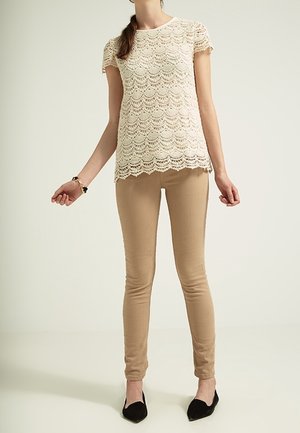 Beige pitsitoppi aaltoilevalla helmalla ja lyhyillä hihoilla, yhdistetty istuviin vaaleanruskeisiin housuihin ja mustiin balleriinoihin. Sileät materiaalit ja monimutkaiset kuviot.