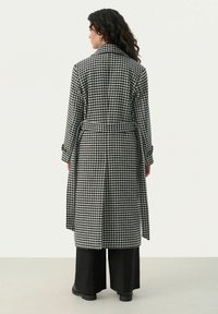 Houndstooth-patterned coat in black and white, featuring a belt, oversized collar, and long sleeves. Paired with wide-leg black trousers.