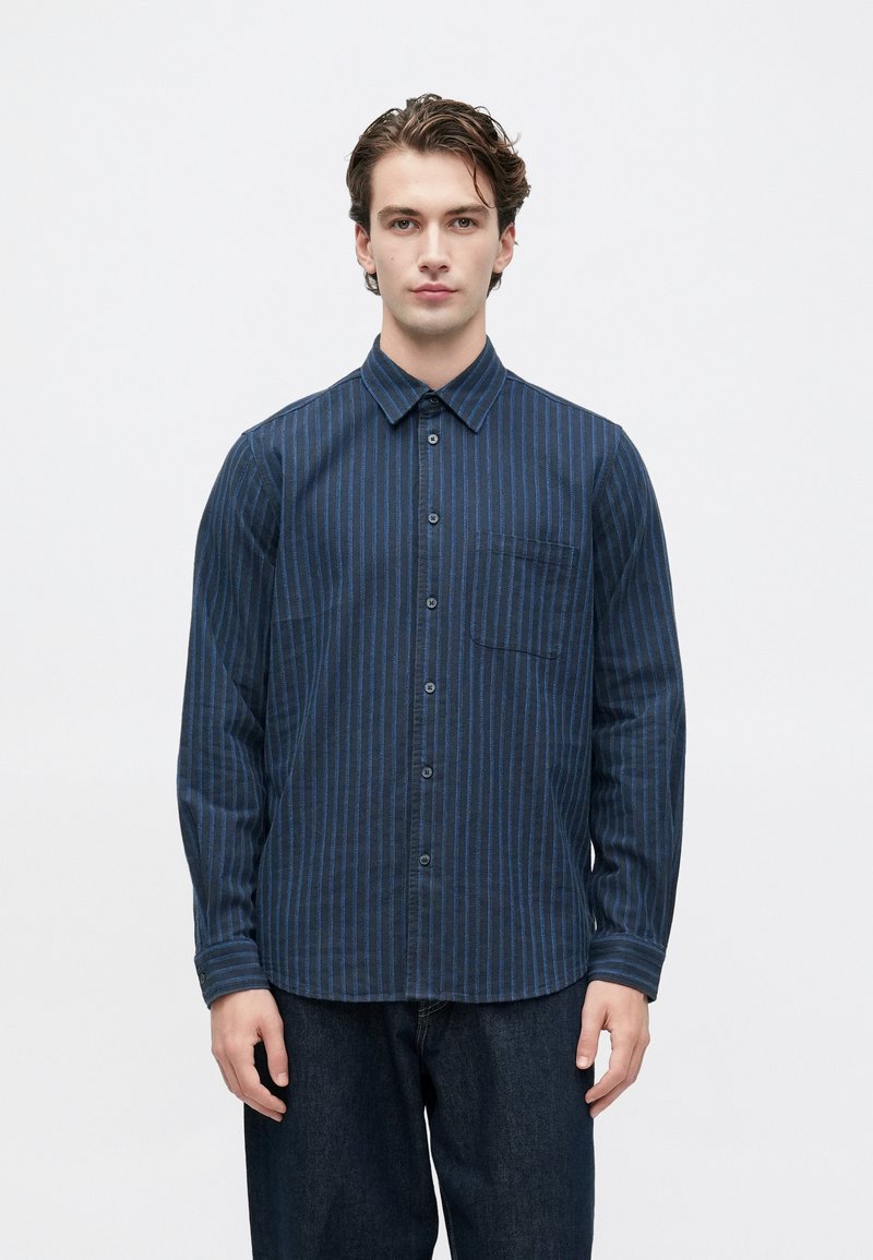 Joven de pie, vistiendo una camisa de manga larga azul marino con botones y rayas verticales, y jeans de un azul oscuro.