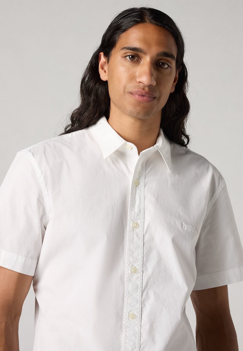 Chemise blanche à manches courtes en coton, avec une texture lisse, dotée d'un col classique et d'un logo discret sur la poitrine.
