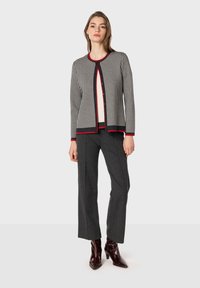 Cardigan a quadre grigi con rifiniture rosse, scollatura rotonda e bottoni, abbinato a pantaloni grigi scuri e stivaletti a punta marroni.