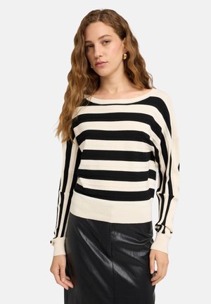 Ung kvinde med bølget hår iført guldringe øreringe, en sort og hvid stribet sweater og et sort lædernederdel, stående mod en hvid baggrund.