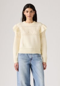 Pull en tricot couleur crème avec des motifs géométriques complexes, épaules froncées et ourlet à côtes. Associé à un jean bleu clair à jambes larges.