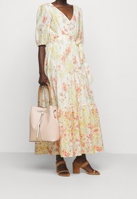 Blommig maxiklänning med V-ringning, puffärmar och volangkjol. Har mönster av rosa och gula blommor mot en ljus bakgrund. Håller en ljusrosa väska.