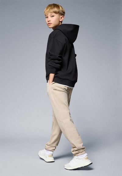 Sweatshirt preto com capuz, calças de moletom bege e ténis brancos com detalhes dourados, exibidos contra um fundo cinza em degradê.