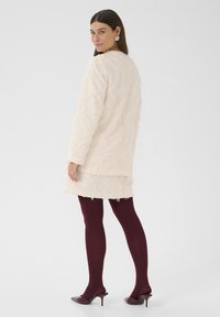 Beige textúrájú kabát, rojtos részletekkel, egy hozzáillő szoknya fölé rétegezve. Sötét burgundi harisnya és hegyes orrú magassarkú cipők teszik teljessé az öltözéket.