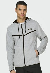 Sudadera gris con cremallera, con una franja de acento negra, cordón ajustable y bolsillos frontales. Tejido suave con una textura lisa.