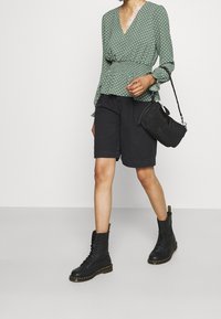 Blusa avvolgente verde a pois con maniche a volants, abbinata a pantaloni corti neri e stivali da combattimento neri con lacci. Anche una borsa a tracolla è inclusa.