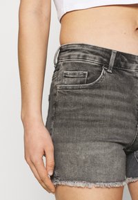 Henkilö, joka käyttää haalistuneita mustia farkkushortsit, joissa on risat reunat, ja valkoista lyhytvartista toppia, käsi lepääntyneenä lonkalla.