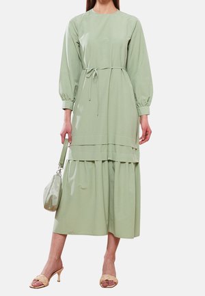 Robe vert clair à manches longues avec une taille à cordon, un design superposé et une texture douce ; accompagnée d'un sac à main assorti et de sandales couleur chair.