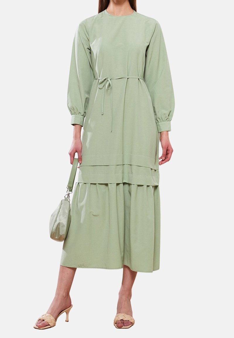 Robe vert clair à manches longues avec une taille à cordon, un design superposé et une texture douce ; accompagnée d'un sac à main assorti et de sandales couleur chair.