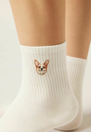 Calzini bianchi alla caviglia a coste, indossati ai piedi, con un piccolo volto di cane marrone e bianco ricamato sul lato esterno vicino alla caviglia.