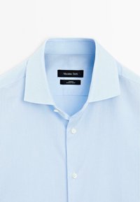 Lichtblauw overhemd met een klassieke kraag, knoopsluiting aan de voorkant en een gestructureerde stof. Etiket leest "Massimo Dutti, Slim Fit, Made in Turkije."