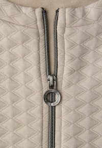 Nahaufnahme eines Metallreißverschlusses mit rundem Zipper an beigem gestepptem Stoff mit Rautenmuster.