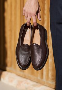 Brązowe mokasyny ze skóry o fakturowanej powierzchni, zaokrąglonym nosku i skórzanej wkładce, trzymane w ręce na tle drewnianym.