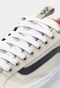 Sneakeren har et beige overdel i suede og mesh, hvite lisser, svart side-stripe og en rød aksent på hælen. Robuste snøringer og teksturerte detaljer.