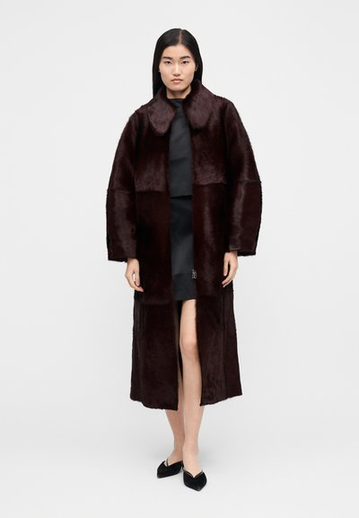 Moteris stovi apsirengusi ilgą tamsiai rudą kailinį paltą, vilkinti juodą suknelę ir juodus batelius su smailiu priekiu, priešais paprastą baltą foną.